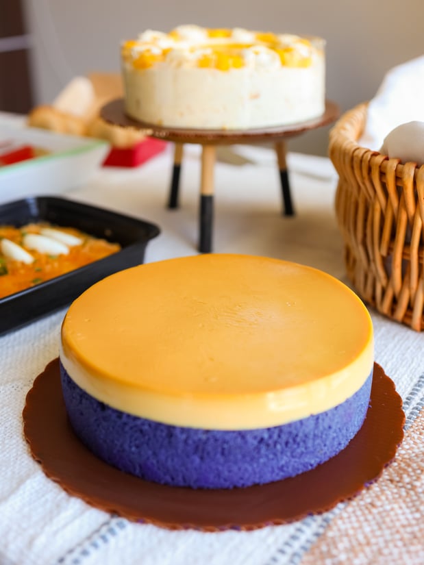 A purple Ube Flan Cake with a bright yellow custard topping sits in the foreground, with a white and yellow Mango Supreme Cake on a small stand behind it.