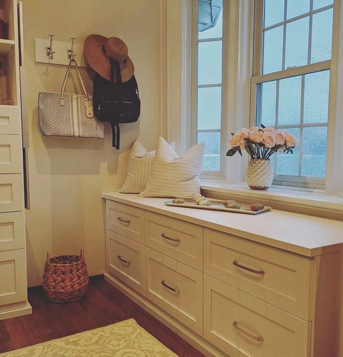 white window seat in closet with window above, pillows, flowers and hooks on wall with hat and purse