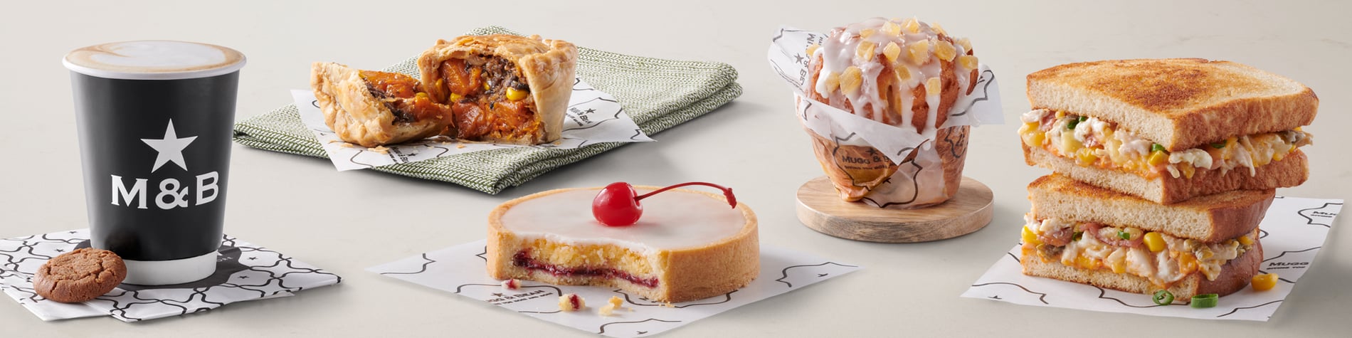 A selection of takeaway meals from the Mugg & Bean On-The-Move menu including a Mexican Chicken Toasted Sandwich, vegetarian Pot Pie, Gingerbread Latte hot drink, freshly baked citrus & ginger muffin, and a slice of Cherry Bakewell Tart.