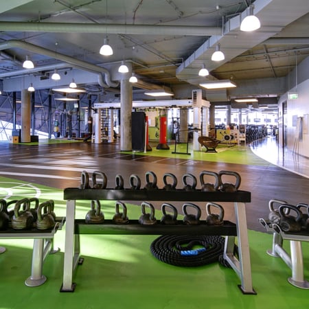 Inside a Fitness First Location, with bright green mats, racks of kettlebells of all sizes, a coiled battle rope and more exercise equipment in the background.