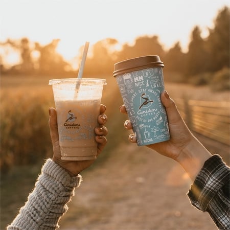 A hand holds up a cup of Iced Coffee in a plastic Caribou Coffee cup and another hand hold up a cup of hot Caribou Coffee in an insulated cup.