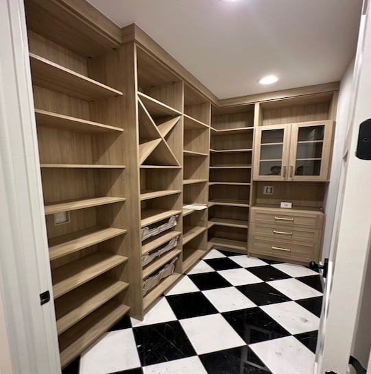 Custom pantry with warm wood-toned shelving, wine rack storage, pull-out baskets, glass-front cabinets, and black-and-white checkered tile flooring.