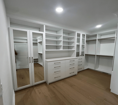 Bright walk-in closet with white built-in shelving, hanging rods, mirrored cabinet doors, and drawer storage, finished with clean shaker-style details and wood flooring.