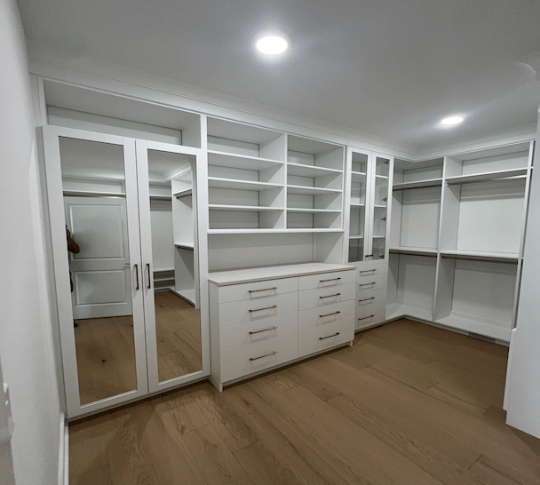 Bright walk-in closet with white built-in shelving, hanging rods, mirrored cabinet doors, and drawer storage, finished with clean shaker-style details and wood flooring.