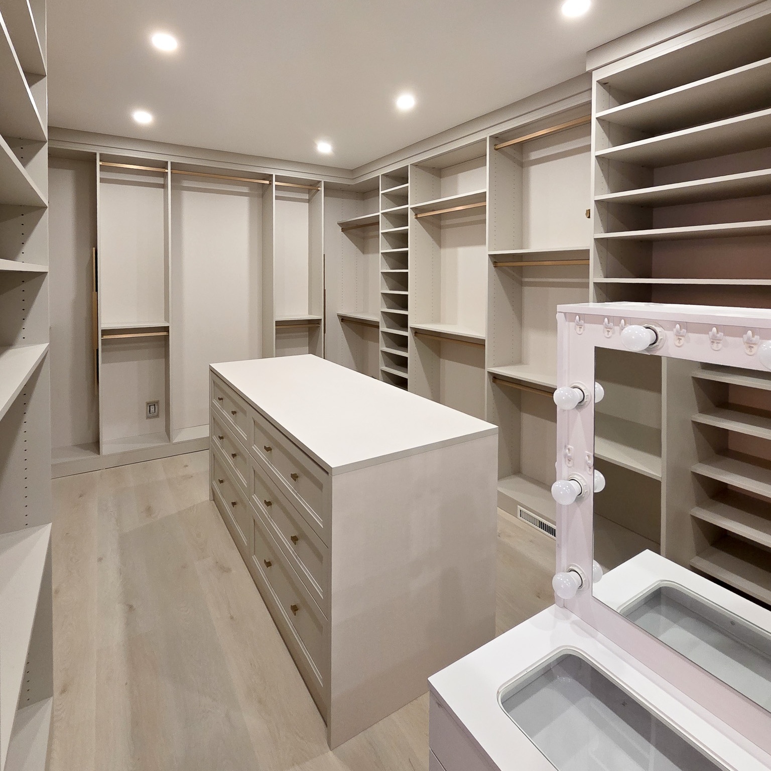 Walk in closet with floor to ceiling storage and a centre island. Designed in a light off white finish with gold hardware.