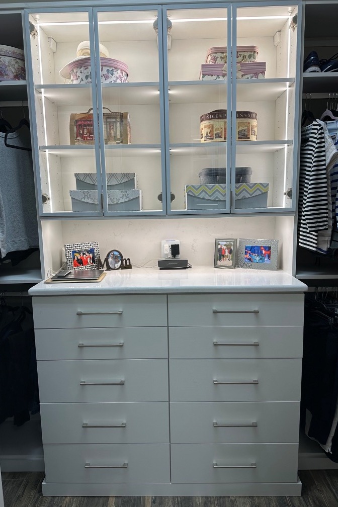 Custom master bedroom closet in Dove White with matte nickel hardware, featuring a hutch-style center drawer bank with enclosed upper cabinets in white aluminum frames, clear glass inserts, and LED strip lighting.