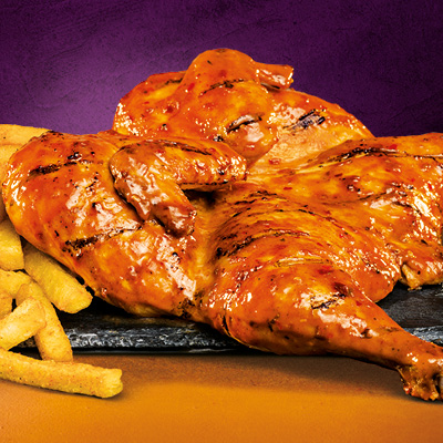 Flame-grilled and full chicken with a serving of chips on the left place on a wooden board against a purple background