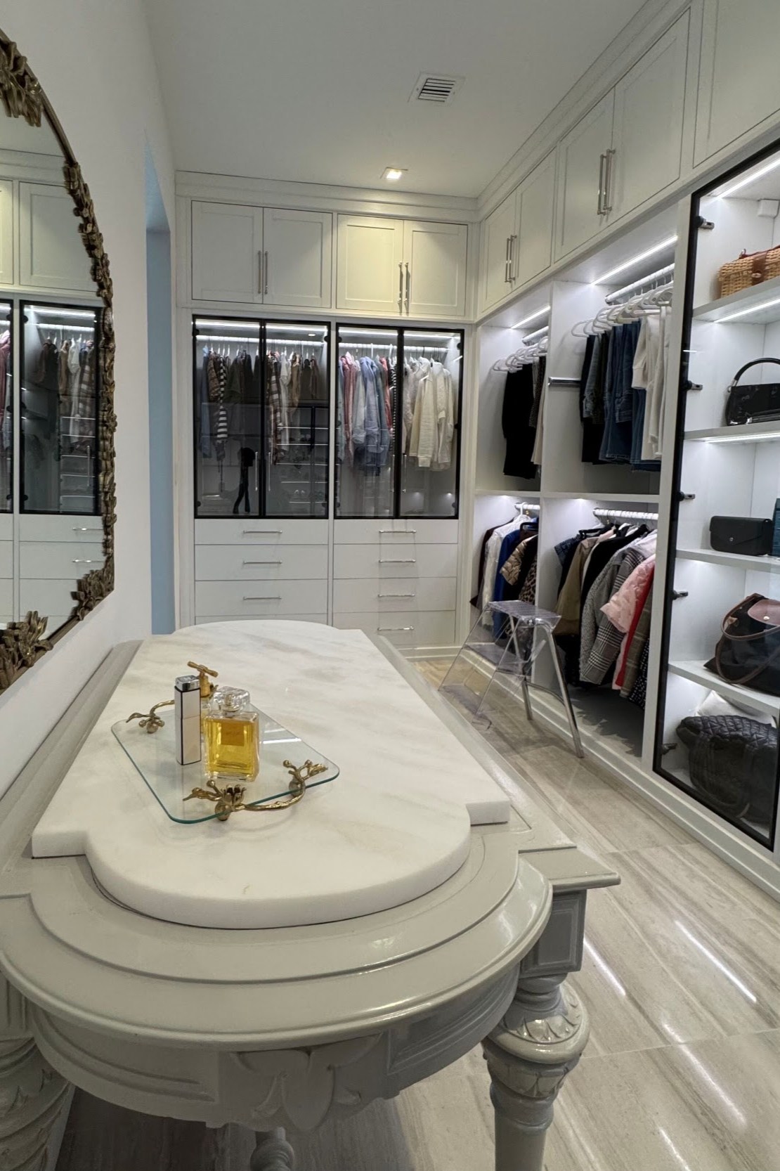 Luxury walk-in closet with white custom cabinetry, glass-front wardrobes, illuminated shelving, hanging storage, and a decorative center island.