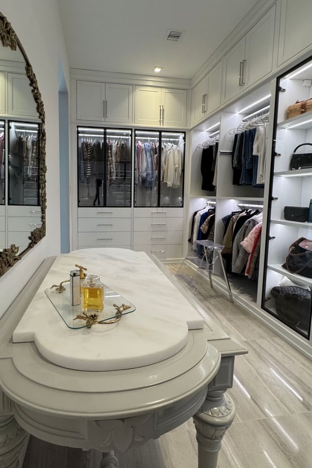 Luxury walk-in closet with white custom cabinetry, glass-front wardrobes, illuminated shelving, hanging storage, and a decorative center island.