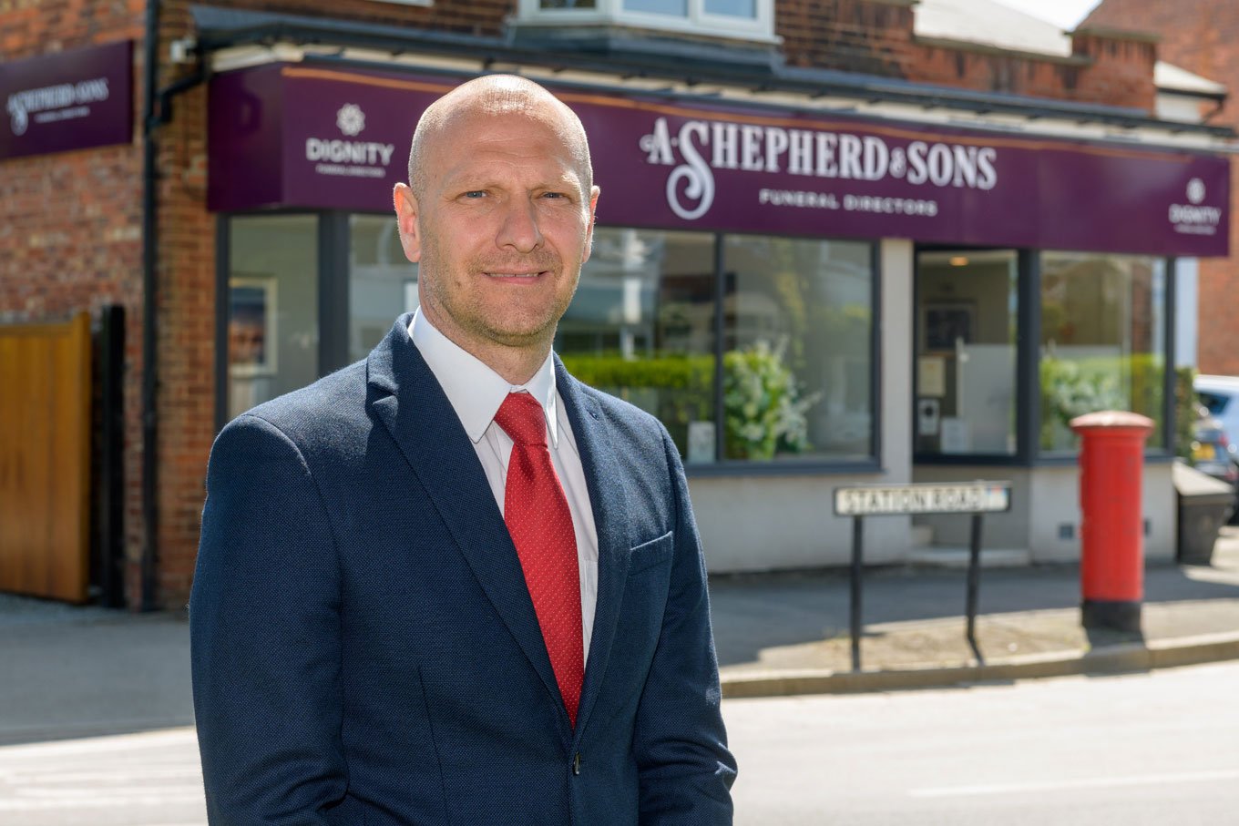 The-Business-Leader-of-A-Shepherd-funeral-directors-stands outside-a-fueral-home-wearing-a-blue-suit-and-a-red-tie