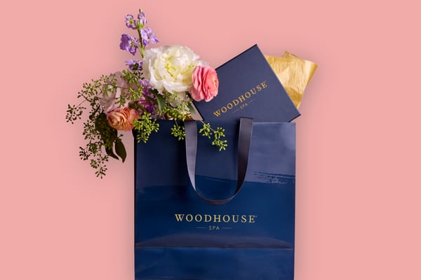 Woodhouse Spa gift bag filled with colorful flowers and a gift card, styled against a soft pink background for a Mother’s Day theme.