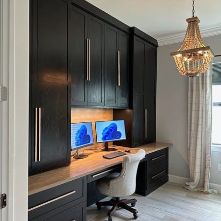 Modern home office with dark custom cabinetry, built-in desk, wood countertop, integrated lighting, and concealed storage.
