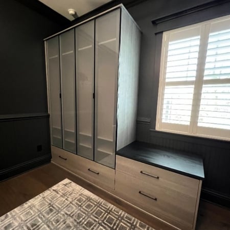 Custom men’s closet in Ash finish with black hardware, aluminum frames with frosted glass inserts, drawers with handles, doors with finger pulls, Everstyle sunglass and jewelry drawers in vegan leather with black trim, and a floating system look with black support legs.