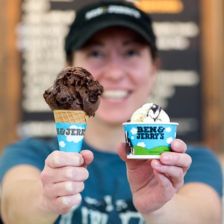 Ben & Jerry's employee holding out a waffle cone with a scoop of chocolate ice cream in the right hand and a cup with a scoop of cookie dough ice cream in the left hand.