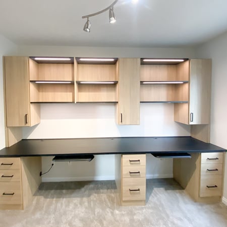 Home office in a natural wood finish with a black countertop. Lighting, open shelves, and closed storage complete this office.