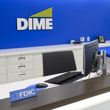 The background of a Dime desk area where the Dime Logo sits in front of a royal blue background and above a white shelving unit and the tops of a computer monitor and computer chair can be seen.