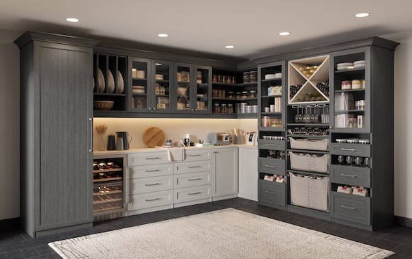 Custom pantry with dark gray cabinets and white lower drawers, featuring glass-front upper shelves filled with jars and dishes, open cubbies with baskets, a wine x, and a small countertop workspace under integrated lighting