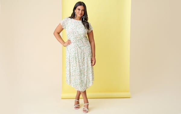 Woman wearing a light green plus size floral midi dress is posing with one hand on her hip in front of a yellow background.