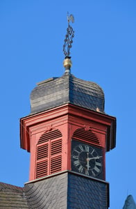Nahaufnahme des Kirchturms. An der Spitze ist eine Uhr zu sehen ´. Die prägenden Farben sind rot und schwarz.