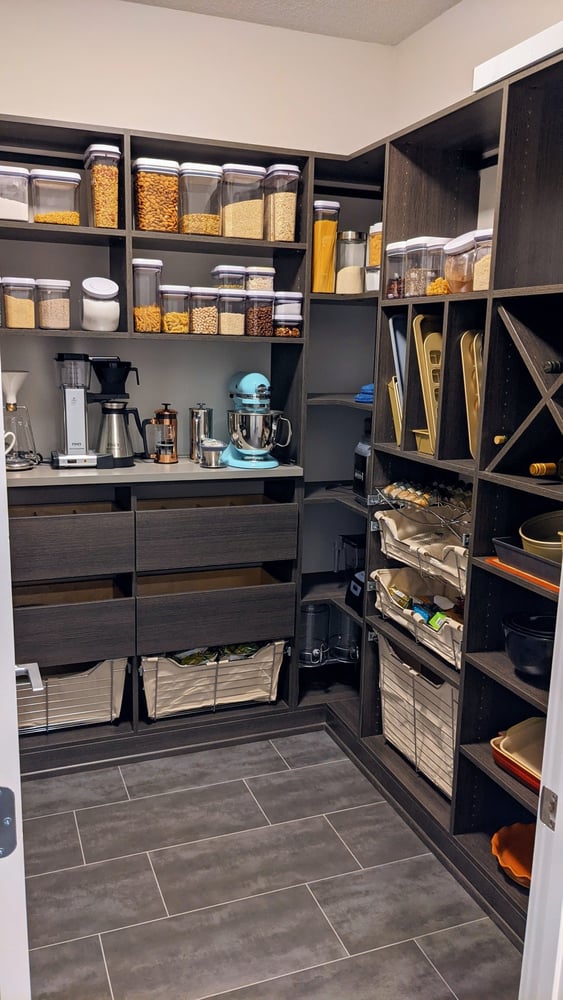 Walk-in pantry designed in a dark wood grain finish. This design included a small countertop for small appliances, There is a combination of pantry drawers and wire baskets with linen lining for produce. There is a combination of horizontal, vertical, corner, and wine shelving.
