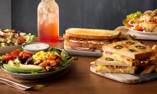 An assortment of generous lunch meals from Mugg & Bean such as toasted sandwiches, salads, and cold drinks on plates on a wooden table with a dark background