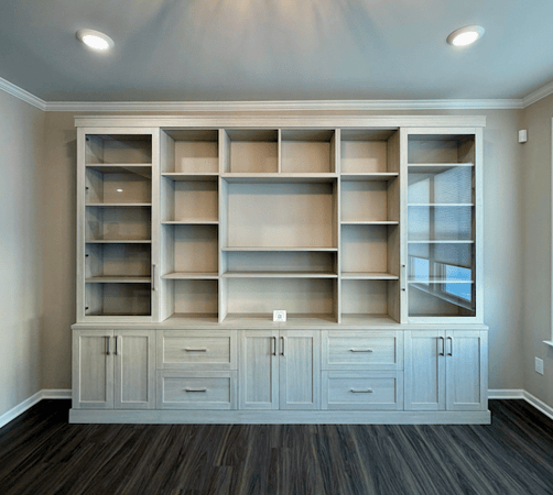 Built-in custom wall bookcase with adjustable open shelving, glass-front side cabinets, and lower drawers and doors in a light gray wood finish with shaker-style detailing by California Closets Michigan in a Milford home.