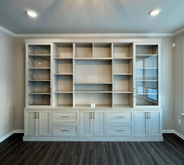 Built-in custom wall bookcase with adjustable open shelving, glass-front side cabinets, and lower drawers and doors in a light gray wood finish with shaker-style detailing by California Closets Michigan in a Milford home.