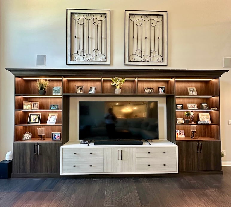 custom wall unit with warm wood shelving and cabinets surrounds a large flat-screen TV. The shelves are softly lit and display framed family photos, small plants, and decorative items.