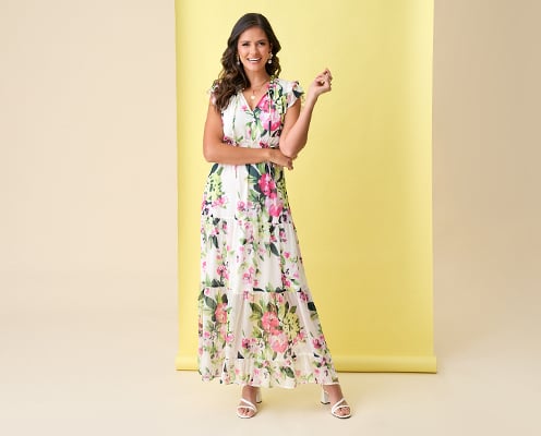 Woman wearing a white floral maxi dress is posing in front of a yellow background.