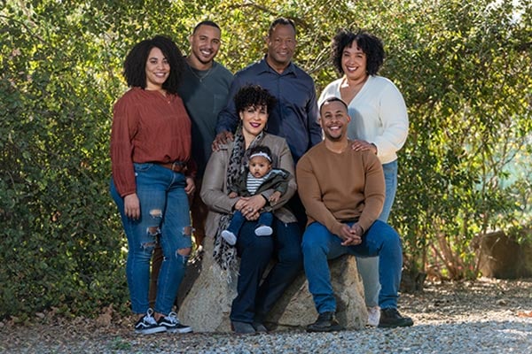 6 smiling family members and a baby sitting in the shade