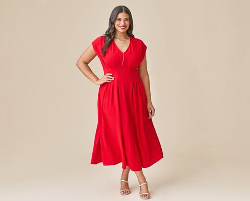 Woman is wearing a plus size red dress and nude heels, and is posing with one hand on her hip in front of a tan background.