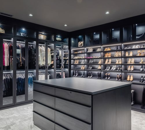 Black walk-in closet with glass door inserts, lighting, center island and slanted shoe shelves.