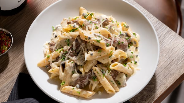 A white bowl filled with penne pasta and pieces of fillet steak in a creamy sauce, topped with grated parmesan and chopped parsley.