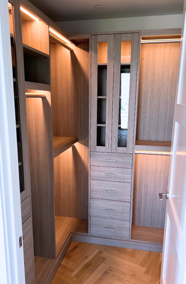 small natural wood beige walk-in closet with double hanging, dress length hanging, LED pole lighting, drawers in middle with silver metal hardware handle, middle shelving, glass doors NYC, New York