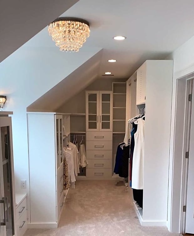 White walk-in closet with sloped ceiling, center cabinet with glass doors above set of drawers