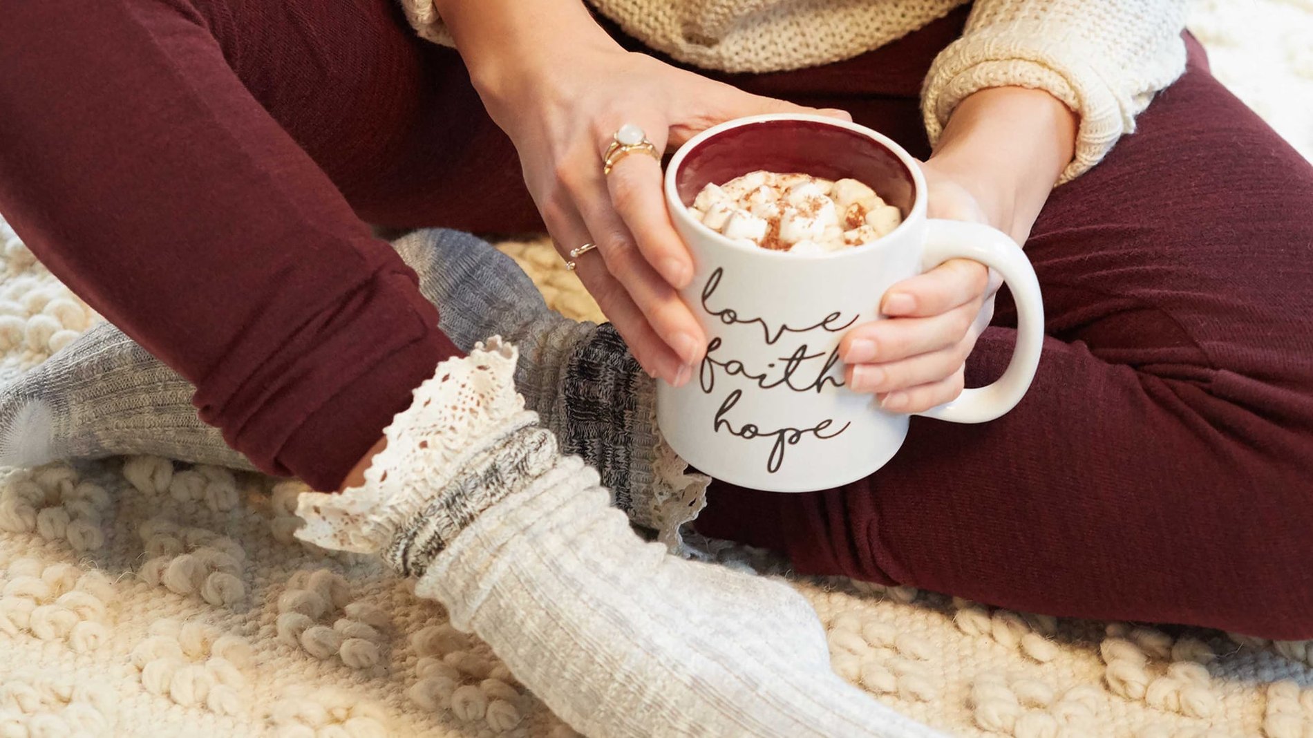 A woman sits on a plush white carpet in comfortable cable knit socks, burgundy leggings and a waffle knit beige sweater, she is holding a mug that says "Love, faith & hope" in cursive.