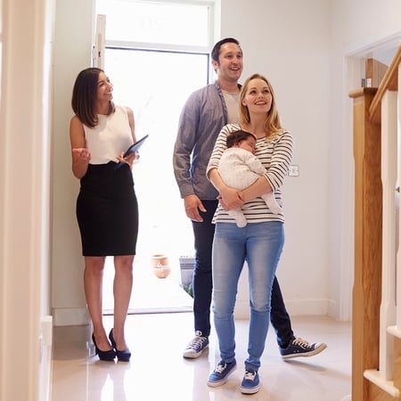A woman in business attire with a man and a woman holding a baby in a house.