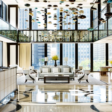 The Lobby of a Langham Hotel with a stunning chandelier, 
a sitting area that includes a plush gray couch and pristine white chairs on an accent rug, behind is a beautiful suburban skyline.