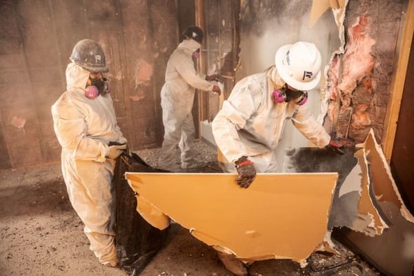 Two pros carry wall debris in
safety gear from a building
with fire and smoke damage.