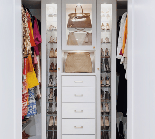 white luxury walk-in closet with drawers in the center and narrow shoe storage on either side with closed glass doors and glass shelving and hanging on either side Westchester New York