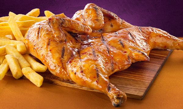 Flame-grilled and full chicken with a serving of chips on the left place on a wooden board against a purple background