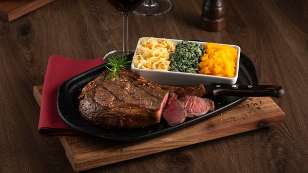 Succulent rib eye on the bone placed on a table with a glass of wine and three side dishes.