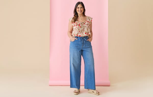 Woman is posing in plus size wide leg jeans and a short sleeve blouse in front of a pink and tan background.
