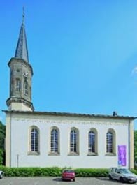 Das Bild zeigt die Außenansicht der Evangelischen Kirche Schwabenheim mit einem weißen Gebäude mit Turm auf der linken Seite. Im Hintergrund vor blauem Himmel.