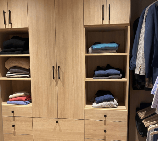 California Closets Westchester NY walk in closet. Natural oak wood color with black hardware for a modern neutral look with open and closed storage space, shelving, double hanging and drawers