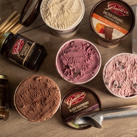 Four different pints of Graeter's Ice cream open with a metal ice cream scoop laid in front, Grater's Butterscotch Dessert Sauce & Bittersweet Dessert Sauce lay on their sides and a stack of disposable ice cream bowls that say "Grater's since 1970" lay on their side.