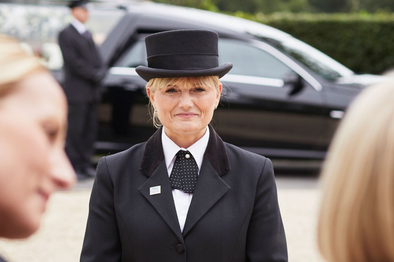 A female funeral director wears a black top hat and gives a compassionate smile to a family.