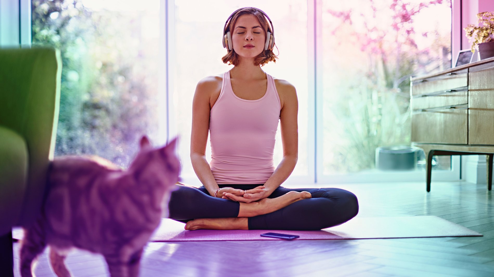 A woman sits on a yoga mat in a living room while wearing large headphones streaming meditation music through her Three Mobile Network on her cell phone that rests on the floor in front of her.