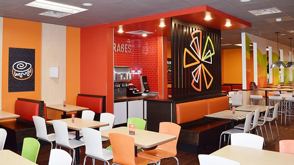 The inside of a Cicis dining room with a mix of brightly colored chairs in front of small tables and plush red booths in against the wall.