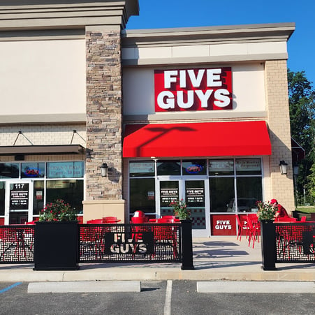 An exterior photograph of the entrance to the Five Guys restaurant at 1629 Publix Way in Stafford, Virginia. The photograph shows the outdoor seating featured at this location, as well as the trademark Five Guys logo sign.